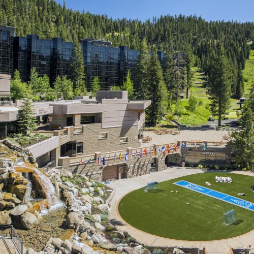 A scenic resort in a green mountainous area features modern architecture, a waterfall, flags, and a circular lawn with a volleyball court.