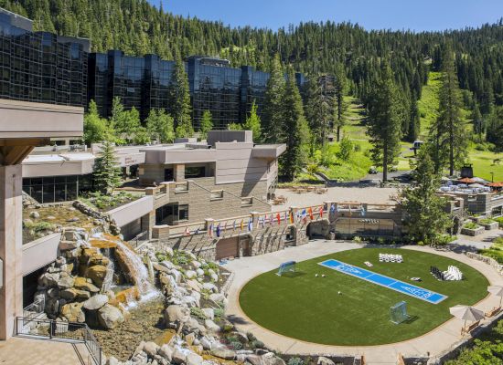 A scenic resort in a green mountainous area features modern architecture, a waterfall, flags, and a circular lawn with a volleyball court.