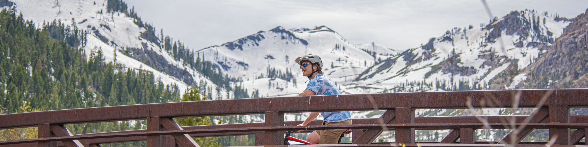 A person is riding a bicycle on a wooden bridge with snow-capped mountains in the background and a partly cloudy sky.