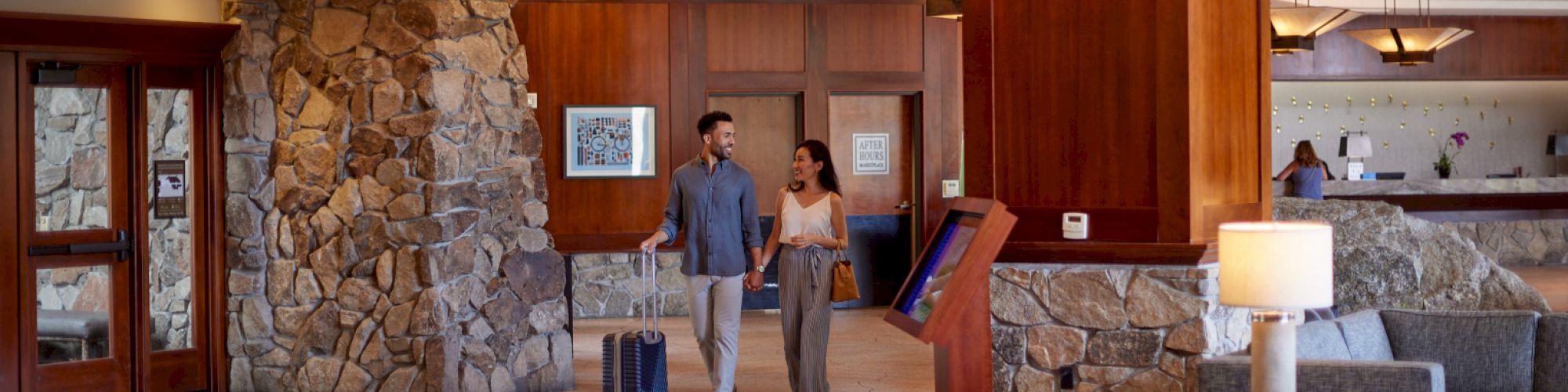 Two people walk through a hotel lobby with suitcases, past a reception desk with staff and seating area.