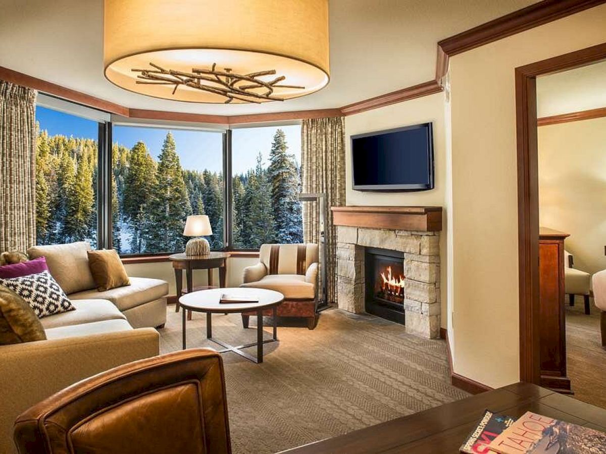 A cozy living room with a fireplace, TV, large windows with forest views, and beige furniture. Adjacent is a bedroom with a lit lamp.