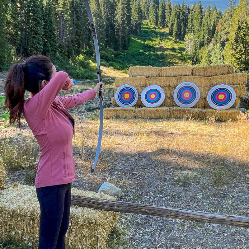 A person is aiming a bow and arrow at a series of four targets in an outdoor archery range surrounded by trees.