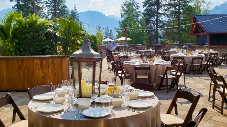 An elegant outdoor dining setup with round tables, chairs, and tableware, surrounded by lush greenery and mountain scenery in the background.