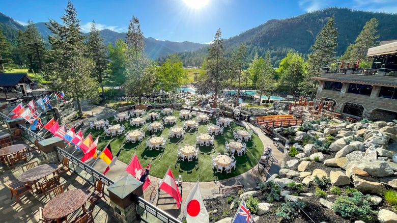An outdoor event venue surrounded by mountains and trees, with circular tables, international flags, and nearby buildings, under a clear sky.