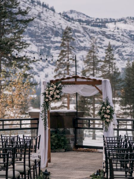 An outdoor wedding setup with chairs facing an arch decorated with flowers, against a snowy, mountainous backdrop.