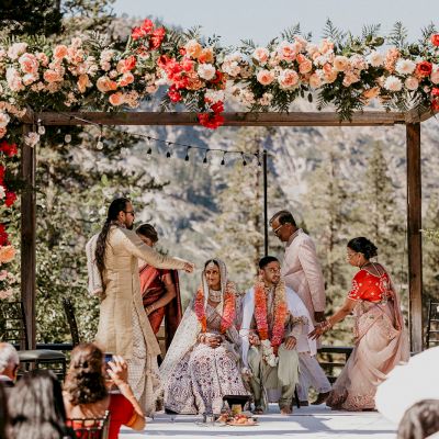 An outdoor wedding ceremony under a floral arch, with a couple in traditional attire seated, surrounded by people, including family members.
