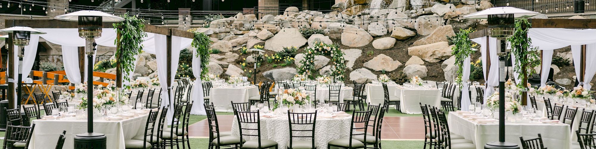 An elegant outdoor event setup with round tables and chairs arranged on a grassy area, surrounded by a backdrop of a building and waterfall.