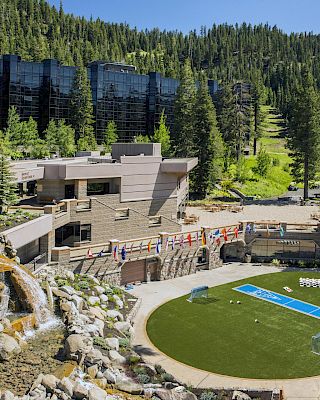 The image shows a modern resort nestled in a forested mountainous area with a waterfall, flags, and recreational areas including a grassy field with a pool.