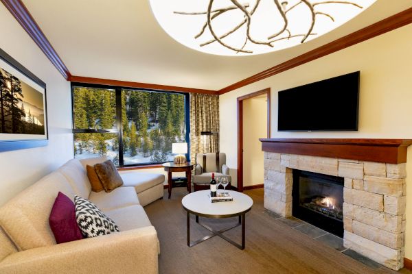 A cozy living room with a beige sofa, a round table, a fireplace, a wall-mounted TV, and a large window with a view of the outdoors.