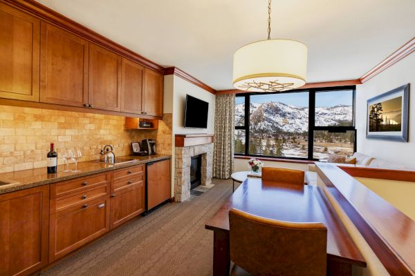 A cozy hotel room with a kitchenette, dining area, fireplace, TV, and a large window offering a picturesque mountain view, with wine on the counter.