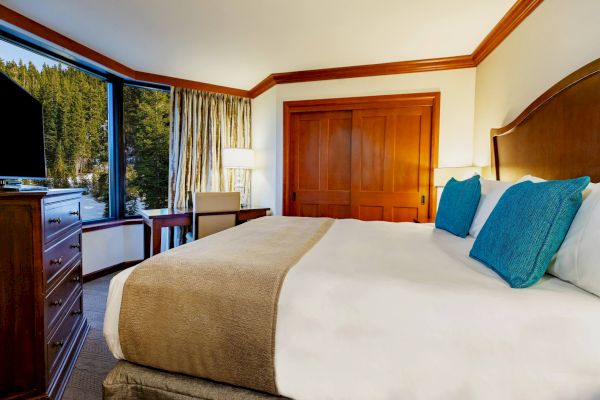 A cozy hotel room features a large bed with blue pillows, a wooden dresser with a TV, a desk, and a large window showcasing a wooded view.