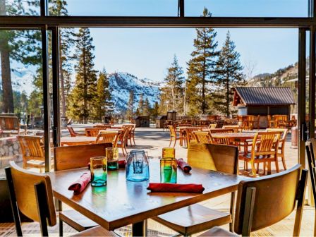 A cozy restaurant with wooden furniture, colorful glasses, set for dining, and a stunning view of trees and snow-covered mountains outside the window.