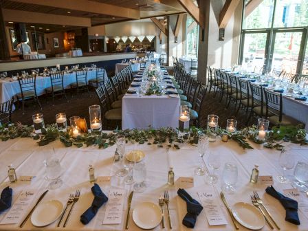 A beautifully set dining area with long tables, greenery, candles, napkins, glassware, and menus, likely prepared for a formal event or wedding.