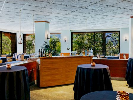 A stylish, well-lit dining area with round tables covered in dark cloths, a bar or reception counter, and large windows overlooking greenery ending the sentence.