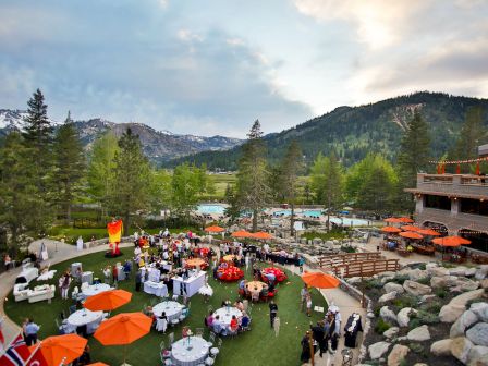An outdoor event with people gathered around tables with orange umbrellas, set against a mountainous backdrop and greenery.