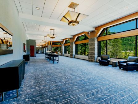 A spacious, modern hallway with large windows, blue carpeting, contemporary lighting fixtures, and black leather armchairs overlooking a scenic view.