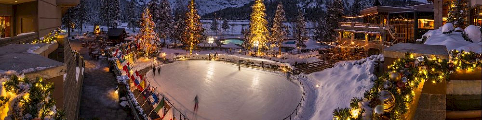 This image shows a festive ice skating rink surrounded by decorated trees and buildings, set in a snowy mountain landscape during dusk.