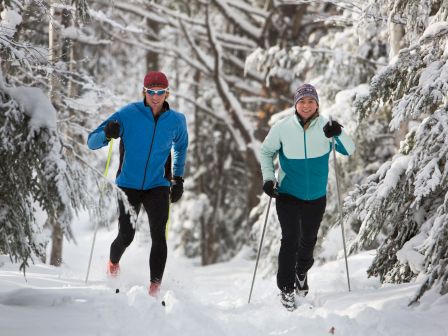 Two people are cross-country skiing through a snowy forest, dressed in winter gear and looking forward.