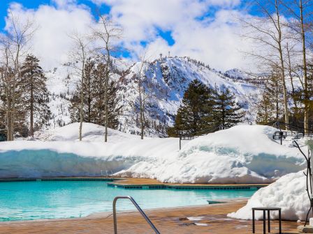 An outdoor pool and hot tub are surrounded by snow-covered landscape with mountains and trees in the background under a blue sky with clouds.