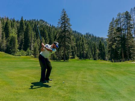 A person is playing golf on a lush green course surrounded by tall trees and rolling hills under a clear blue sky.