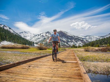 A person rides a bike on a wooden path through a scenic landscape with snowy mountains, green trees, and a blue sky with wispy clouds, ending the sentence.