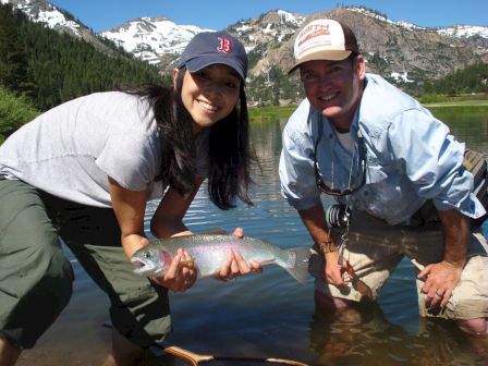 A woman and a man are posing by a river holding a caught fish, with snowy mountains in the background and both smiling.
