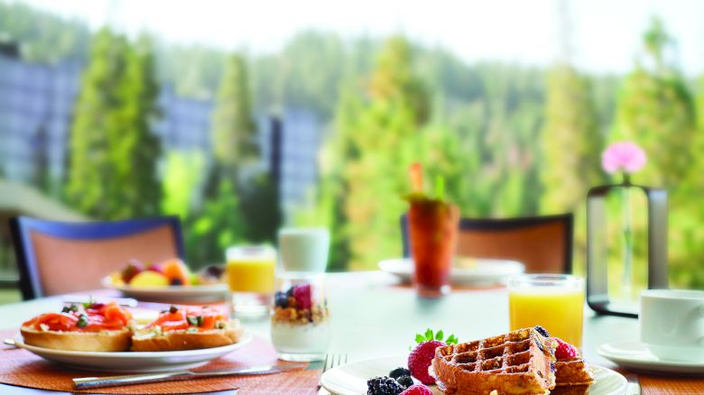 A table set with a breakfast including waffles, fruit, yogurt, and juice, with a scenic outdoor view in the background.