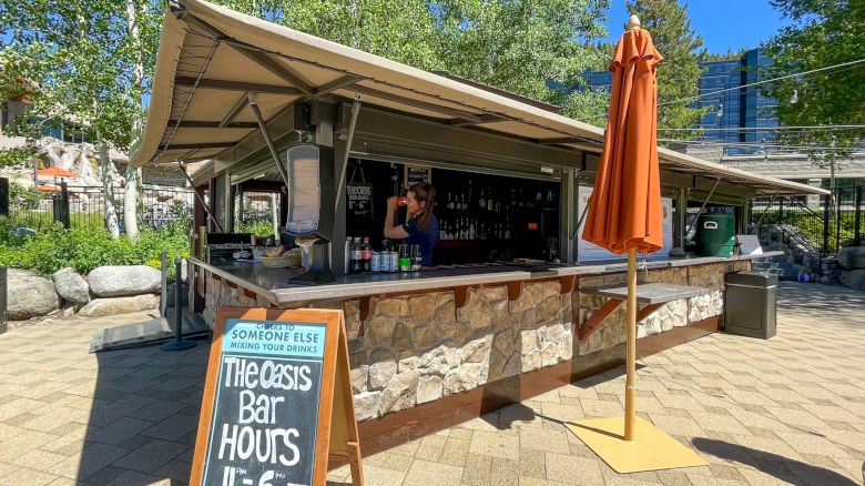 An outdoor bar with a stone facade and awning, featuring a chalkboard sign with hours, various drinks, and an orange umbrella. The end.