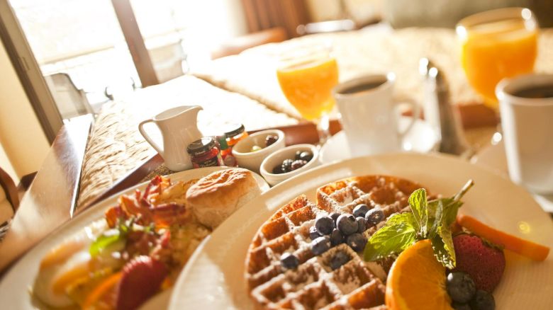 A breakfast spread featuring waffles with blueberries and powdered sugar, orange slices, strawberries, coffee, juice, and various small condiments.