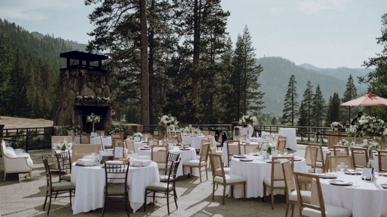 An outdoor event setup with round tables adorned with white tablecloths and floral arrangements, surrounded by a scenic forest and mountains background.