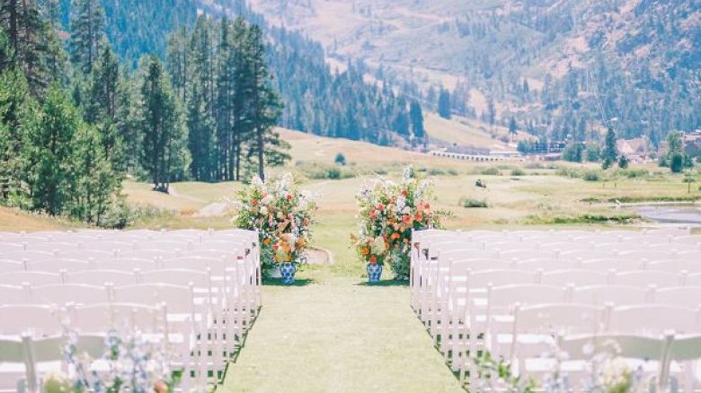 An outdoor wedding setup with rows of white chairs on either side of a grassy aisle, decorated with flowers, mountains, and trees in the background.