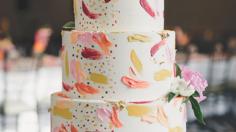 A three-tiered cake with colorful brushstroke decorations and floral accents on top and around, placed on an ornate silver cake stand.