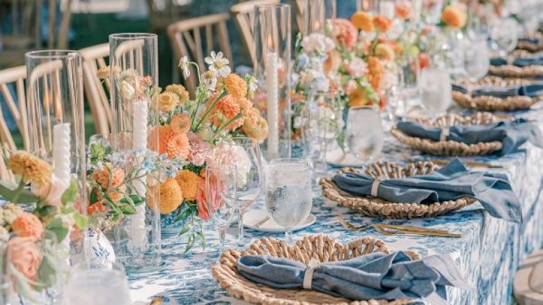 A long, elegantly set outdoor table with floral arrangements, candles, woven placemats, and blue napkins is ready for a gathering or celebration.