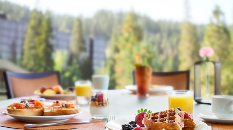 A breakfast table with waffles, juice, fruit, and a parfait, set against a blurry forest view.