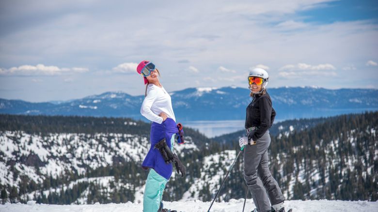 Two people in ski gear posing on a snow-covered mountain with scenic views of trees and mountains in the background.