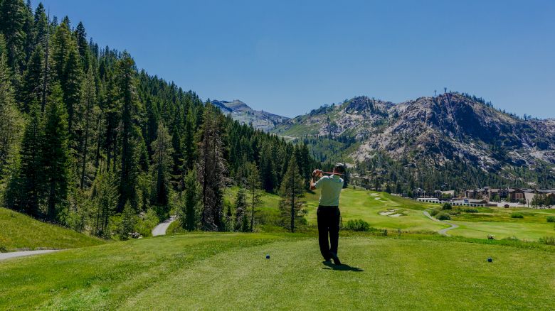 A golfer is teeing off on a lush green golf course surrounded by mountains and pine trees under a clear blue sky.