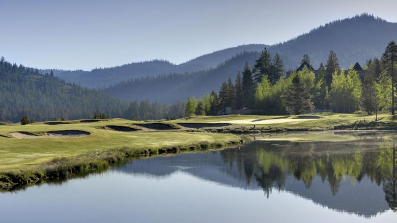 A picturesque golf course with a water hazard, surrounded by forested hills and mountains, and a reflection of trees in the water.