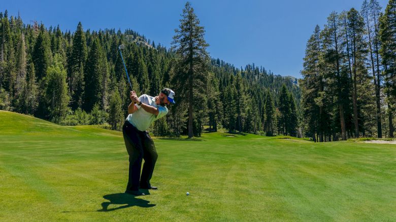 A golfer is mid-swing on a lush, green course surrounded by tall trees and scenic mountains under a clear, blue sky.