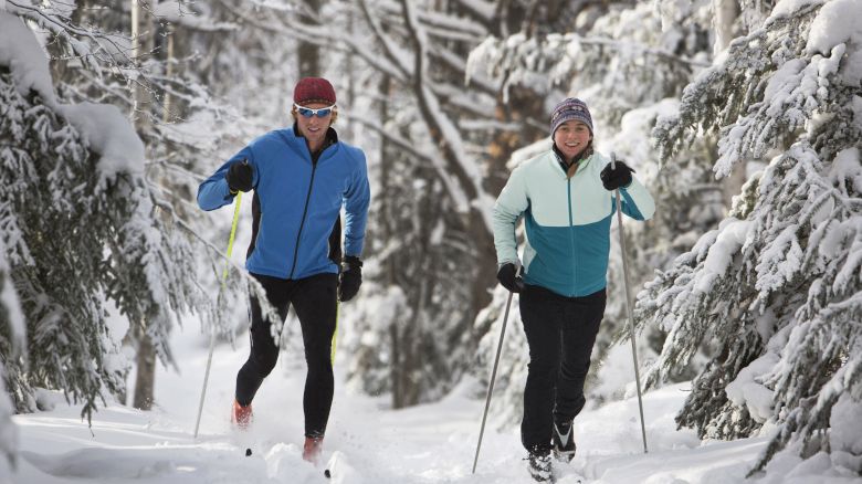 Two people cross-country skiing on a snow-covered trail in a forest, dressed in winter gear and smiling.