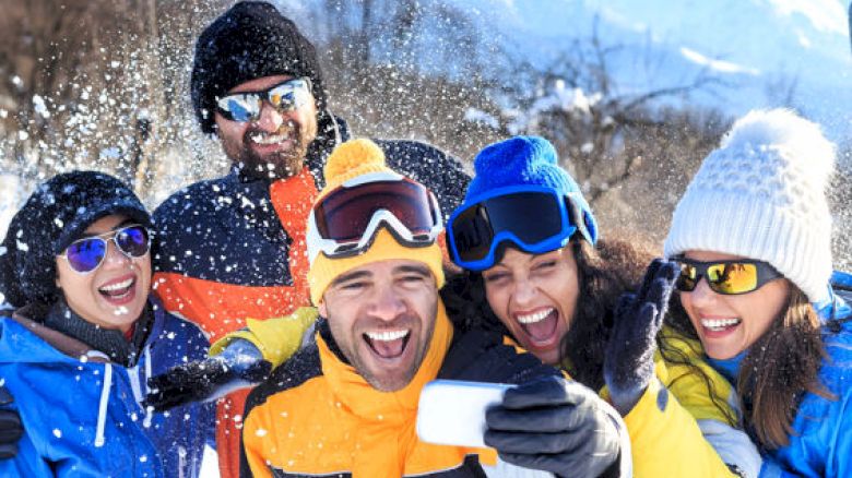 A group of friends in winter gear, having fun in a snowy mountain setting, taking a selfie and smiling cheerfully under the bright sun.