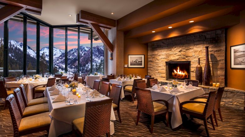 A cozy dining room with a large fireplace, elegantly set tables, and large windows showcasing a snowy mountain view at sunset.