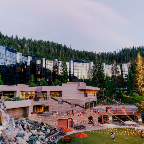 This image depicts a picturesque resort nestled in a forested mountainous area, featuring modern buildings with glass exteriors, a waterfall, and string lights.