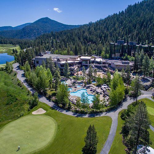 An aerial view of a resort with a central pool area, surrounded by lush greenery, golf course sections, and mountains in the background.