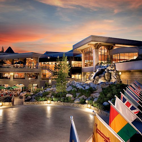 A resort with modern architectural buildings, national flags, and a statue, set against a backdrop of a vibrant sunset sky and natural surroundings.