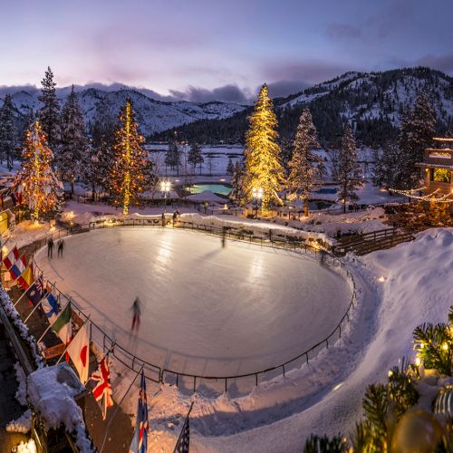 A beautifully illuminated outdoor ice skating rink surrounded by snowy scenery and festive decorations, with mountains in the background.