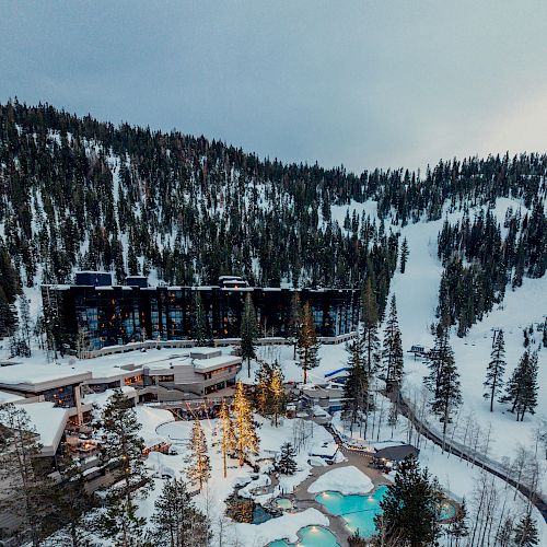 A snow-covered resort nestled in pine trees features a main building and several outdoor pools surrounded by a mountainous landscape.