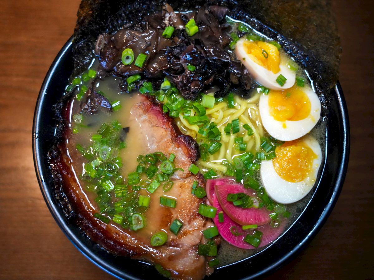 A bowl of ramen featuring slices of pork, boiled eggs, noodles, green onions, pickled radish, and mushrooms in a rich broth on a wooden surface.