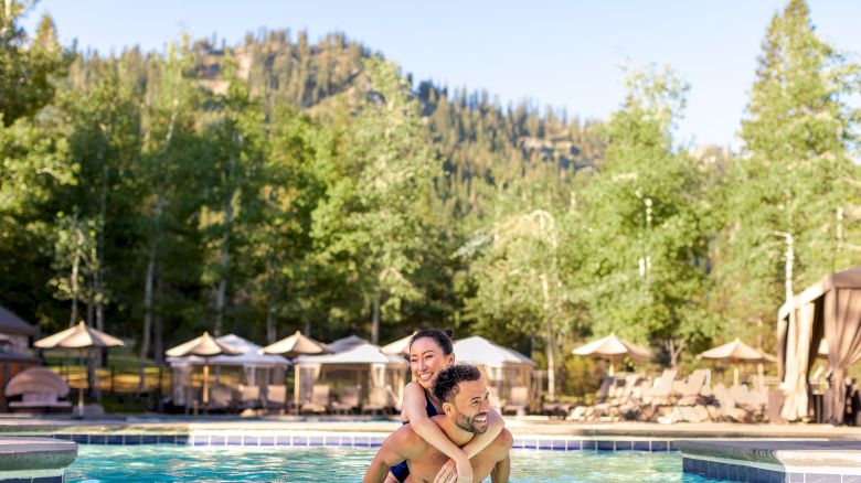 A couple is enjoying a playful moment in a pool, set against a backdrop of trees and cabanas with a mountainous landscape in the background.