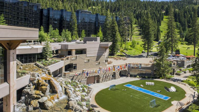 The image shows a modern resort nestled in a forested mountainous area, featuring buildings, a fountain, and a turf area with flags and seating.