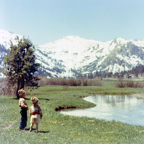 Two young children stand near a river, looking at snow-covered mountains in the background, surrounded by greenery and trees.